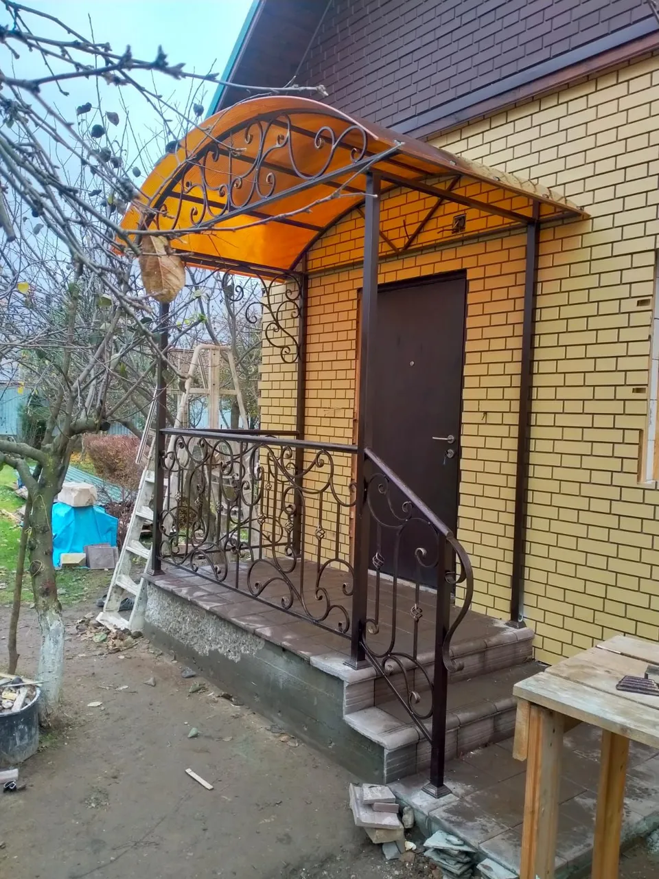 Крыльцо с коваными перилами и арочным навесом из янтарного поликарбоната